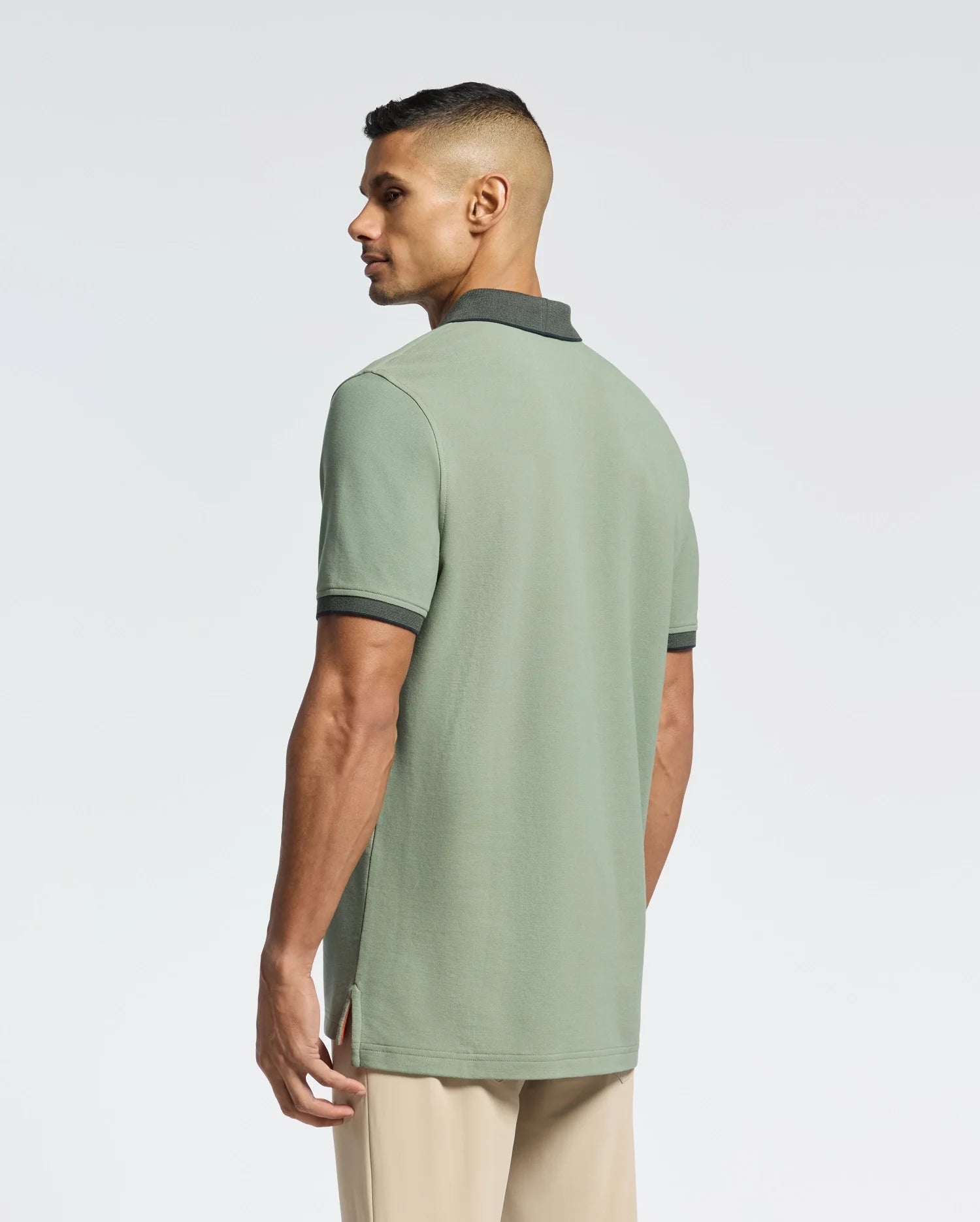 A person with short hair, their back to the camera, wears a Psycho Bunny light green pique polo featuring dark green trim, beige pants, and an embroidered bunny on the pocket. The backdrop is plain and light-colored.