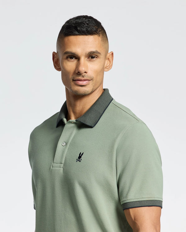 A man in a Psycho Bunny MENS JONAS STRIPE COLLAR PIQUE POLO, light green with a dark collar and embroidered bunny logo, looks at the camera against a white background.