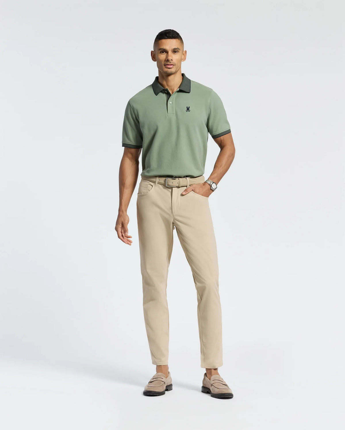 A man stands against a plain background in a Psycho Bunny MENS JONAS STRIPE COLLAR PIQUE POLO - B6K583F200, paired with beige pants and suede loafers. Sporting a short haircut and a watch on his left wrist, he maintains a relaxed posture and neutral expression.