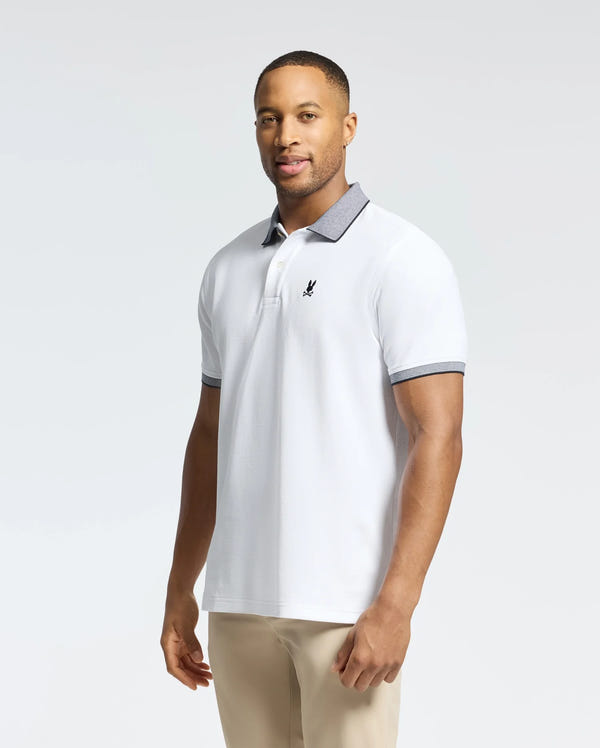 A man wears the Psycho Bunny MENS JONAS STRIPE COLLAR PIQUE POLO - B6K583F200, featuring a white micro-stripe design with gray collar and cuffs. His light pants complement the polo's small black emblem on the left, indicating its premium Pima cotton quality, as he looks neutrally at the camera.