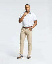 A person in a Psycho Bunny white Pima cotton Jonas Stripe Collar Pique Polo, beige trousers, and brown sneakers stands on a white background. They wear a watch on their left wrist with one hand touching their chin.