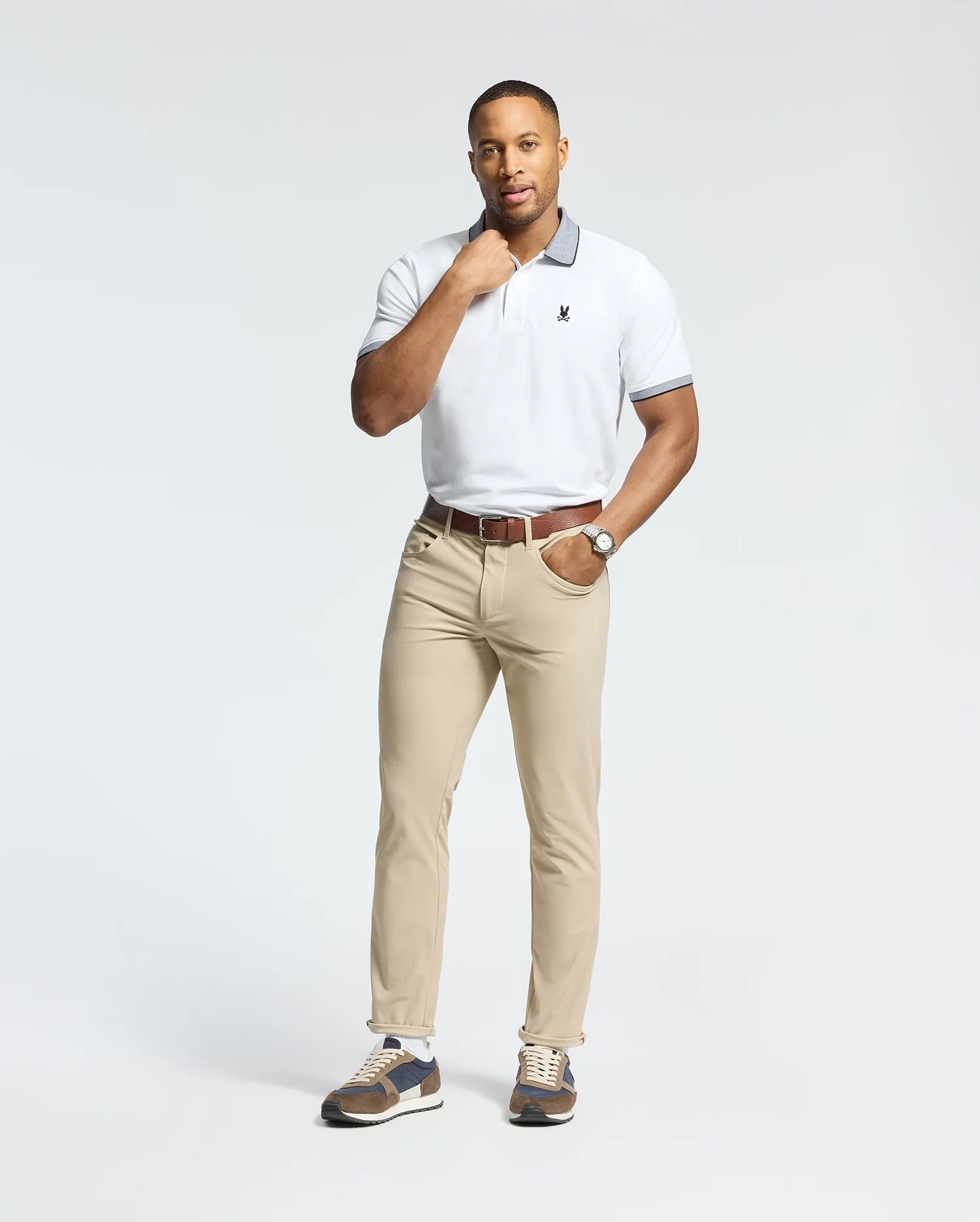A person in a Psycho Bunny white Pima cotton Jonas Stripe Collar Pique Polo, beige trousers, and brown sneakers stands on a white background. They wear a watch on their left wrist with one hand touching their chin.
