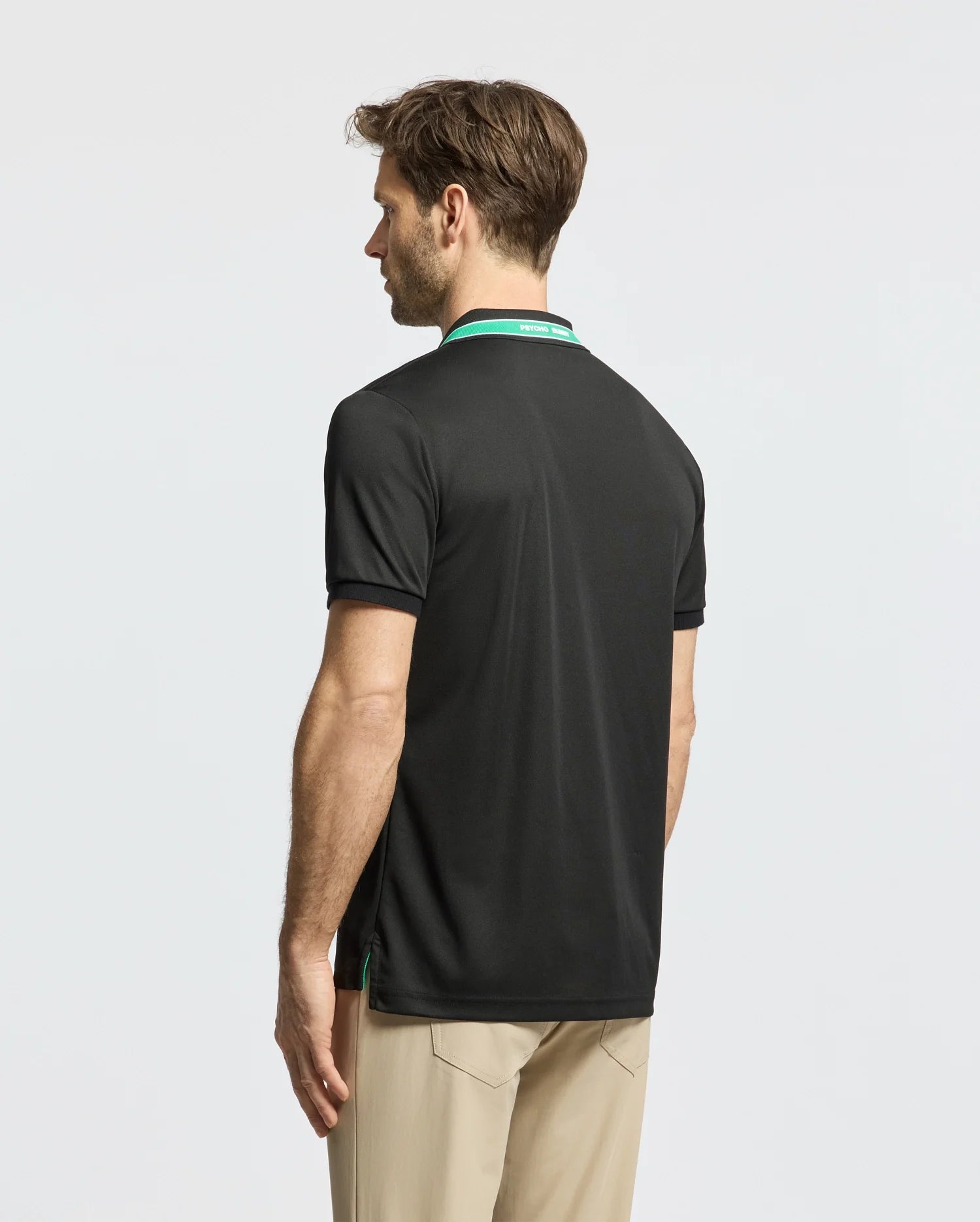 A man wearing the Psycho Bunny MENS FORREST SPORT PIQUE POLO - B6K660F200, black with green accents, paired with beige pants, stands turned sideways against a plain white background. The slim-fit polo is made from moisture-wicking fabric for both comfort and style.