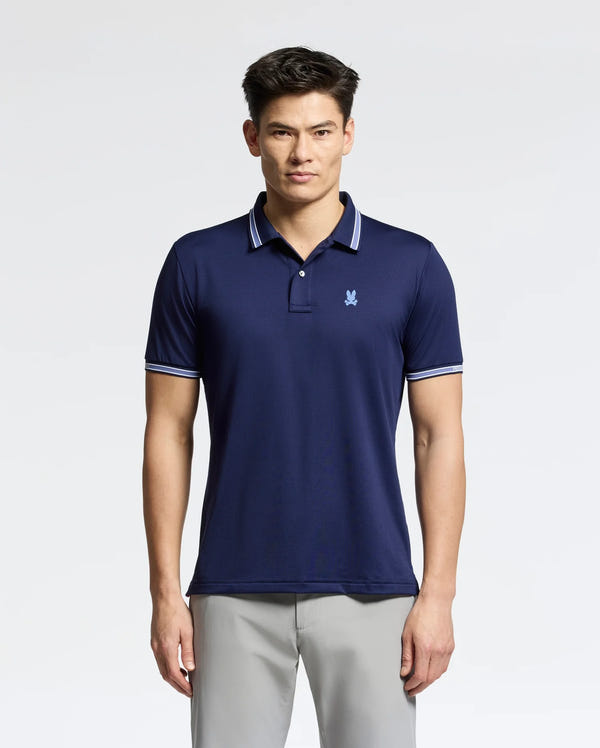 A man in a navy blue Psycho Bunny MENS FORREST SPORT PIQUE POLO - B6K660F200, featuring white-trimmed collar and sleeves, exudes style with its slim fit and small chest logo. The breathable fabric complements his light gray pants as he poses against a plain light background.