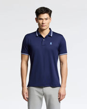 A man in a navy blue Psycho Bunny MENS FORREST SPORT PIQUE POLO - B6K660F200, featuring white-trimmed collar and sleeves, exudes style with its slim fit and small chest logo. The breathable fabric complements his light gray pants as he poses against a plain light background.