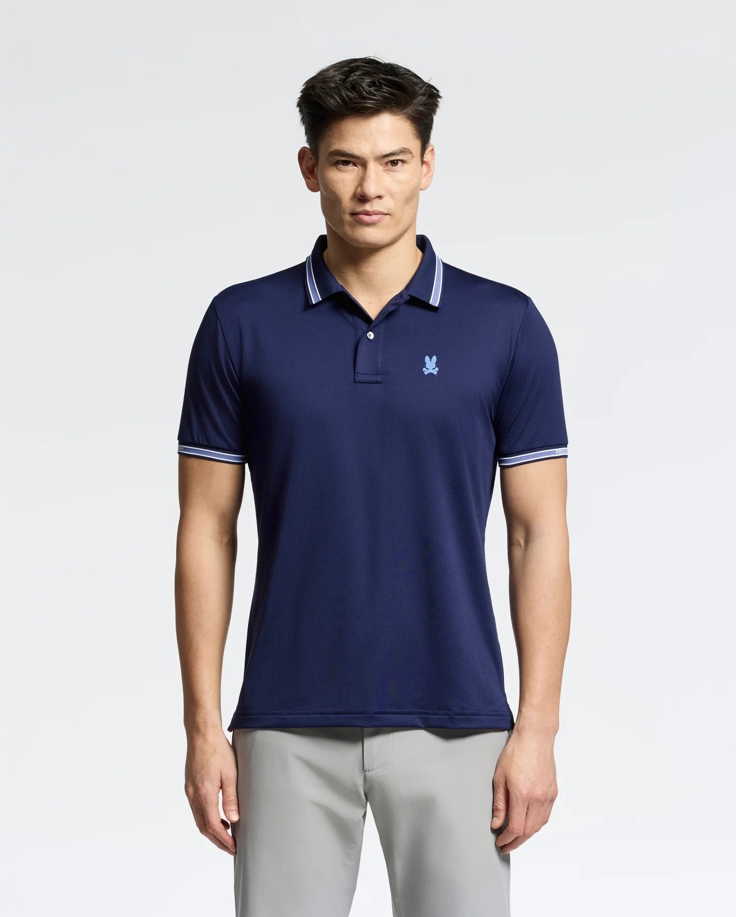 A man in a navy blue Psycho Bunny MENS FORREST SPORT PIQUE POLO - B6K660F200, featuring white-trimmed collar and sleeves, exudes style with its slim fit and small chest logo. The breathable fabric complements his light gray pants as he poses against a plain light background.
