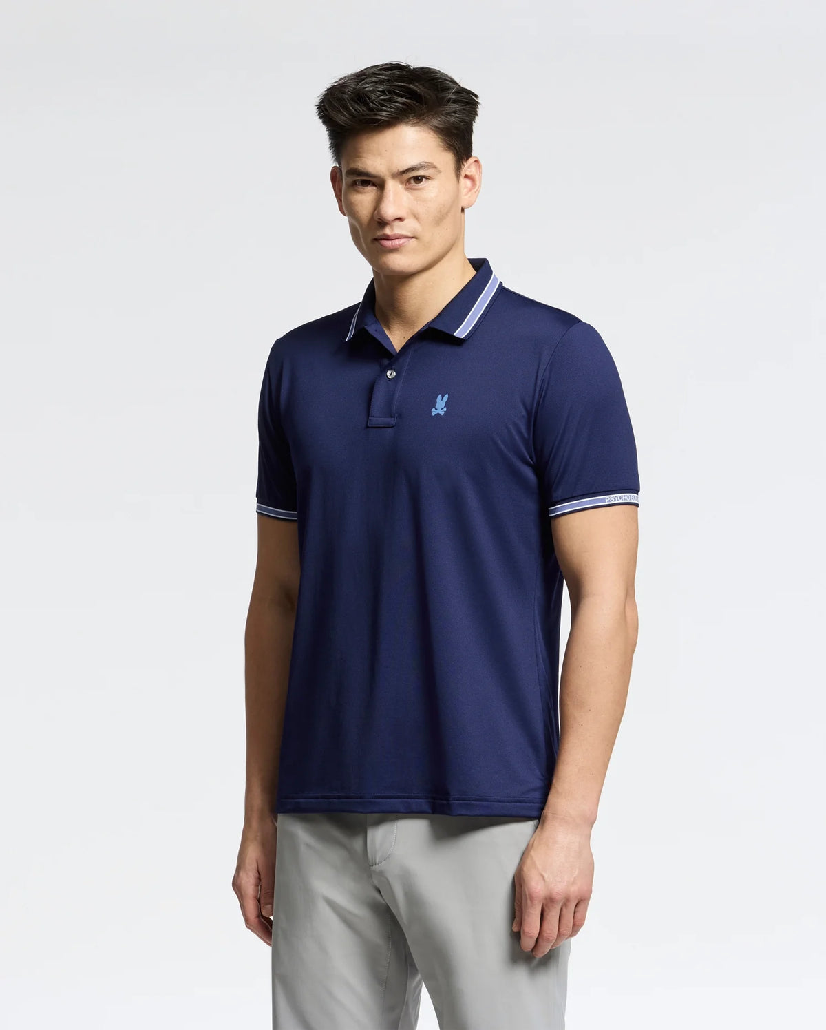 A person wearing the Psycho Bunny navy MENS FORREST SPORT PIQUE POLO - B6K660F200 in breathable fabric with a light blue logo and white-trimmed collar and sleeves. The slim fit accentuates their stature. They have short, dark hair, standing confidently against a plain white background.