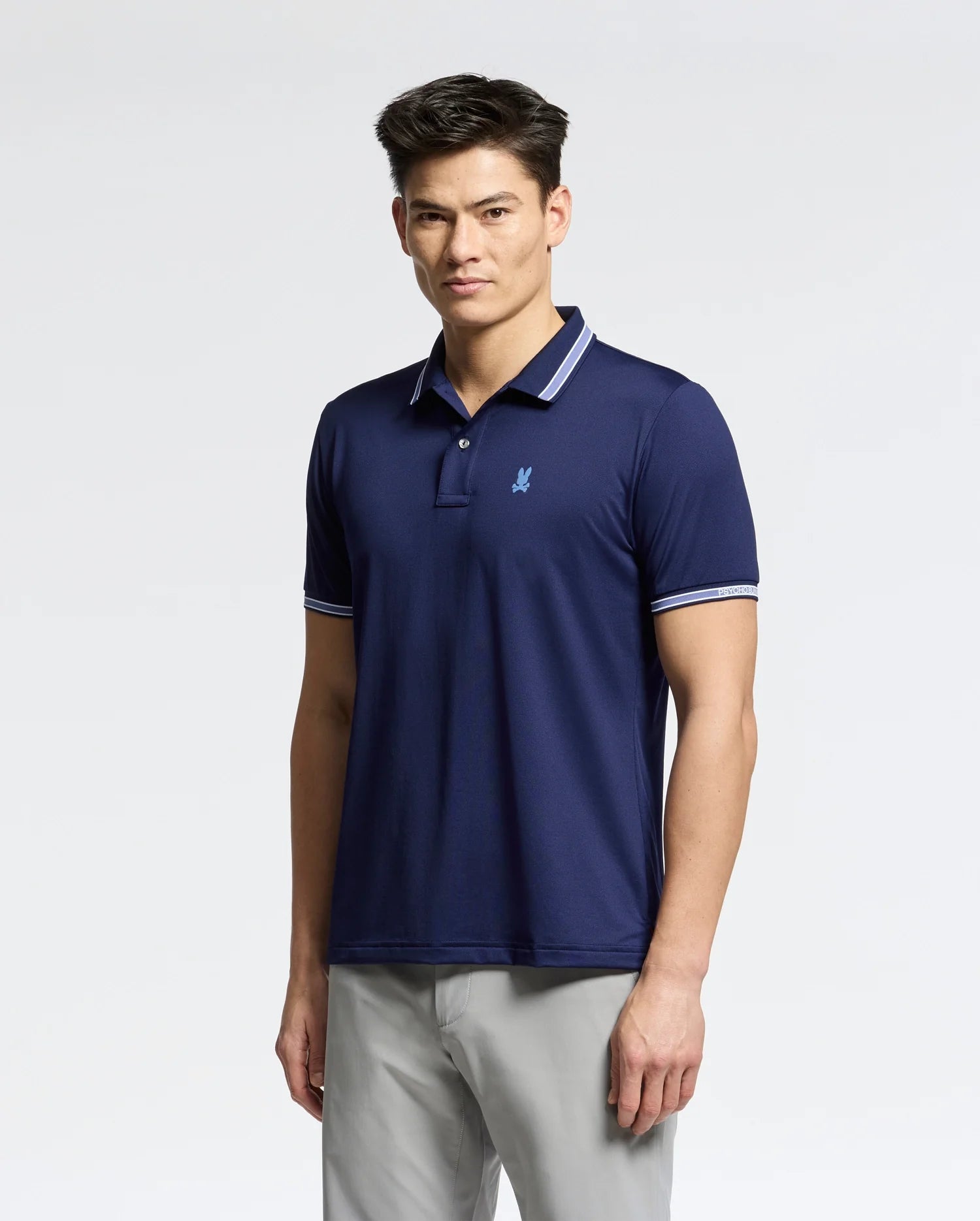 A person wearing the Psycho Bunny navy MENS FORREST SPORT PIQUE POLO - B6K660F200 in breathable fabric with a light blue logo and white-trimmed collar and sleeves. The slim fit accentuates their stature. They have short, dark hair, standing confidently against a plain white background.