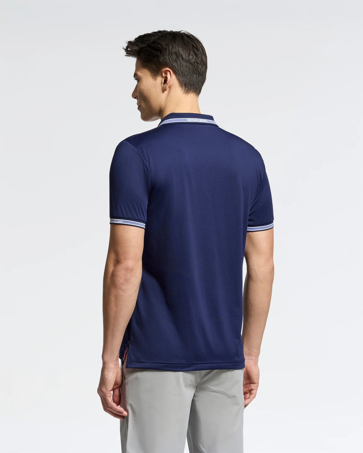 A person with short dark hair is standing and facing away, wearing Psycho Bunny's MENS FORREST SPORT PIQUE POLO (B6K660F200), a slim fit navy blue shirt in breathable fabric with white trim on the collar and sleeves. They pair it with light gray pants against a plain white background.