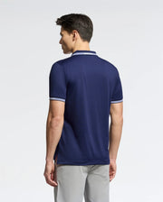 A person with short dark hair is standing and facing away, wearing Psycho Bunny's MENS FORREST SPORT PIQUE POLO (B6K660F200), a slim fit navy blue shirt in breathable fabric with white trim on the collar and sleeves. They pair it with light gray pants against a plain white background.