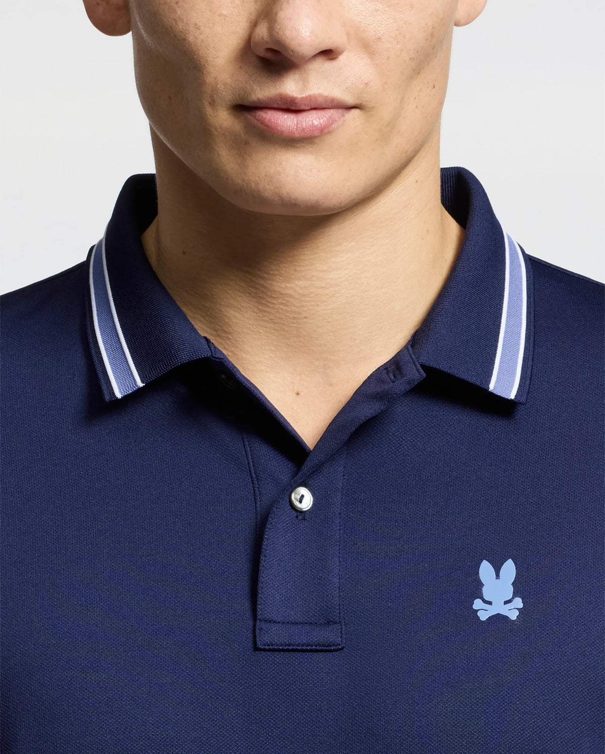 A person is seen from the shoulders up, wearing the Psycho Bunny MENS FORREST SPORT PIQUE POLO - B6K660F200, a slim fit navy shirt featuring a small light blue bunny logo and an embroidered skull on the chest. Its breathable fabric provides comfort against the light background.