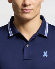 A person is seen from the shoulders up, wearing the Psycho Bunny MENS FORREST SPORT PIQUE POLO - B6K660F200, a slim fit navy shirt featuring a small light blue bunny logo and an embroidered skull on the chest. Its breathable fabric provides comfort against the light background.