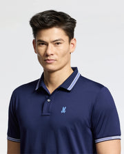 A person with short dark hair wears the Psycho Bunny MENS FORREST SPORT PIQUE POLO - B6K660F200 in blue, featuring a small embroidered logo. Crafted from breathable fabric, they stand against a plain light background, looking directly at the camera with a neutral expression.
