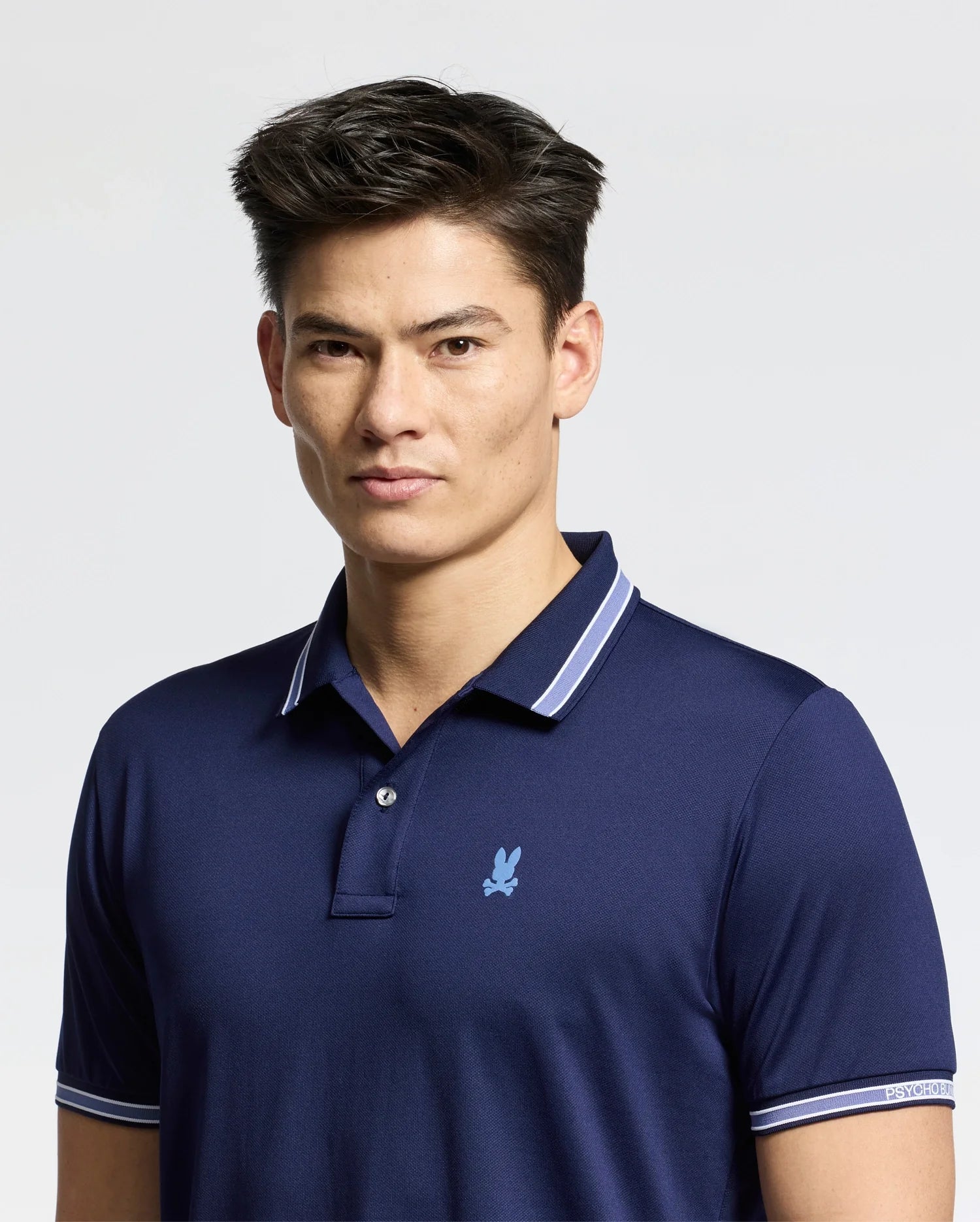 A person with short dark hair wears the Psycho Bunny MENS FORREST SPORT PIQUE POLO - B6K660F200 in blue, featuring a small embroidered logo. Crafted from breathable fabric, they stand against a plain light background, looking directly at the camera with a neutral expression.