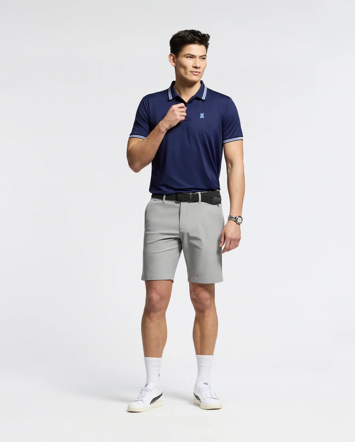 A person confidently poses against a white backdrop wearing Psycho Bunny's navy MENS FORREST SPORT PIQUE POLO, light gray shorts, black belt, and white sneakers. The breathable fabric enhances their style as they adjust the collar with one hand while showcasing a watch on the other.