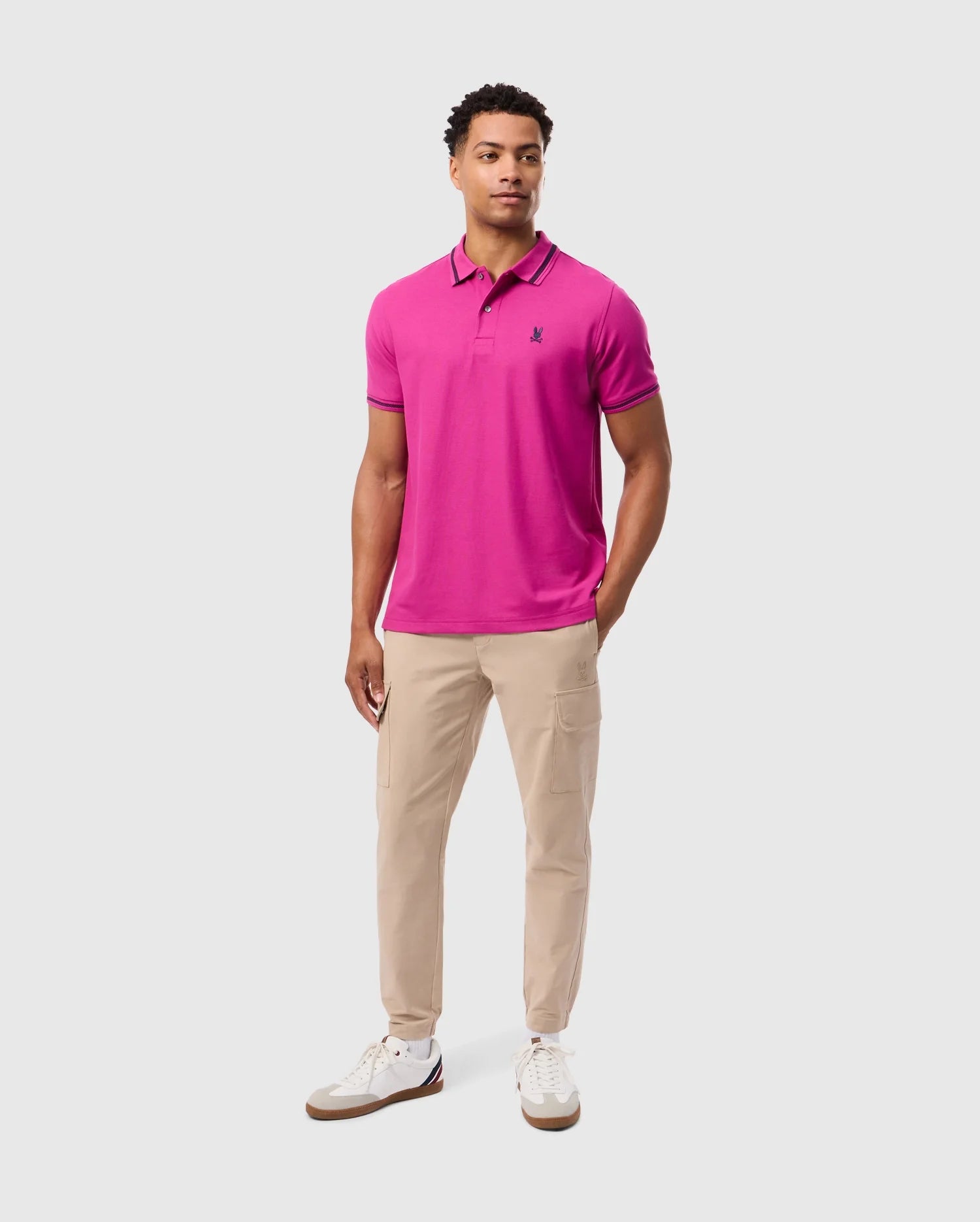 A man stands wearing a vibrant pink Psycho Bunny MENS RIVIERA PIQUE POLO SHIRT (B6K640C200), featuring an embroidered Bunny appliqué, paired with beige cargo pants and white sneakers against a plain background.