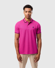 A man stands wearing a vibrant pink Psycho Bunny MENS RIVIERA PIQUE POLO SHIRT - B6K640C200, made from soft Pima cotton and showcasing an embroidered Bunny appliqué on the chest. He has short curly hair and pairs the polo with beige pants against a plain light gray background.