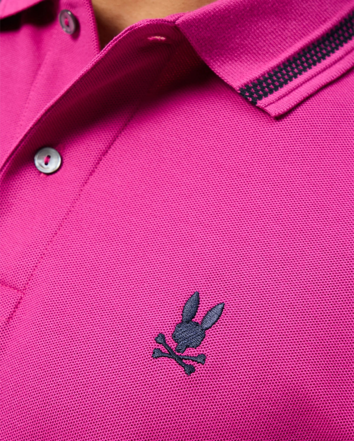 This close-up showcases the Psycho Bunny Men's Riviera Pique Polo Shirt - B6K640C200 in magenta, made from soft Pima cotton. The design includes a black and white checkered pattern on the collar with an embroidered Bunny appliqué of a rabbit's head above crossbones on the chest.