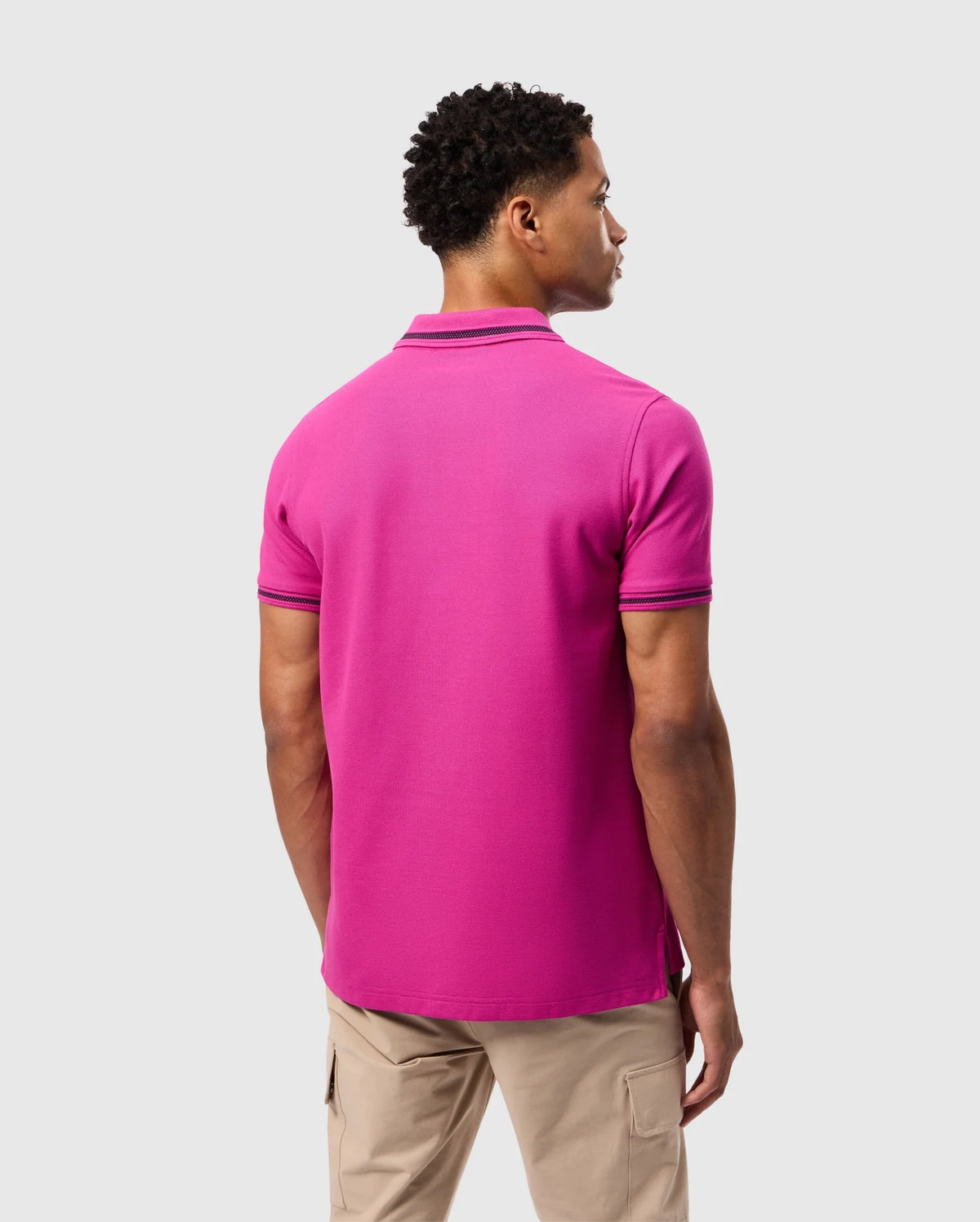 A person stands with their back to the camera, wearing a vibrant pink Psycho Bunny Riviera Pique polo shirt, crafted from soft Pima cotton and paired with beige pants. The sleek design offers a subtle nod to an intricate embroidered Bunny appliqué, set against a plain grey background.