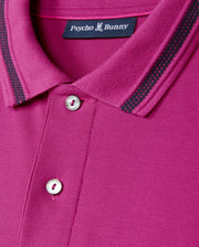 Close-up of the MENS RIVIERA PIQUE POLO SHIRT by Psycho Bunny in a vibrant magenta Pima cotton, featuring a collar with black and gray striped details. The shirt boasts two buttons, an embroidered Bunny appliqué, and a label at the collar that reads "Psycho Bunny" with a small bunny logo.