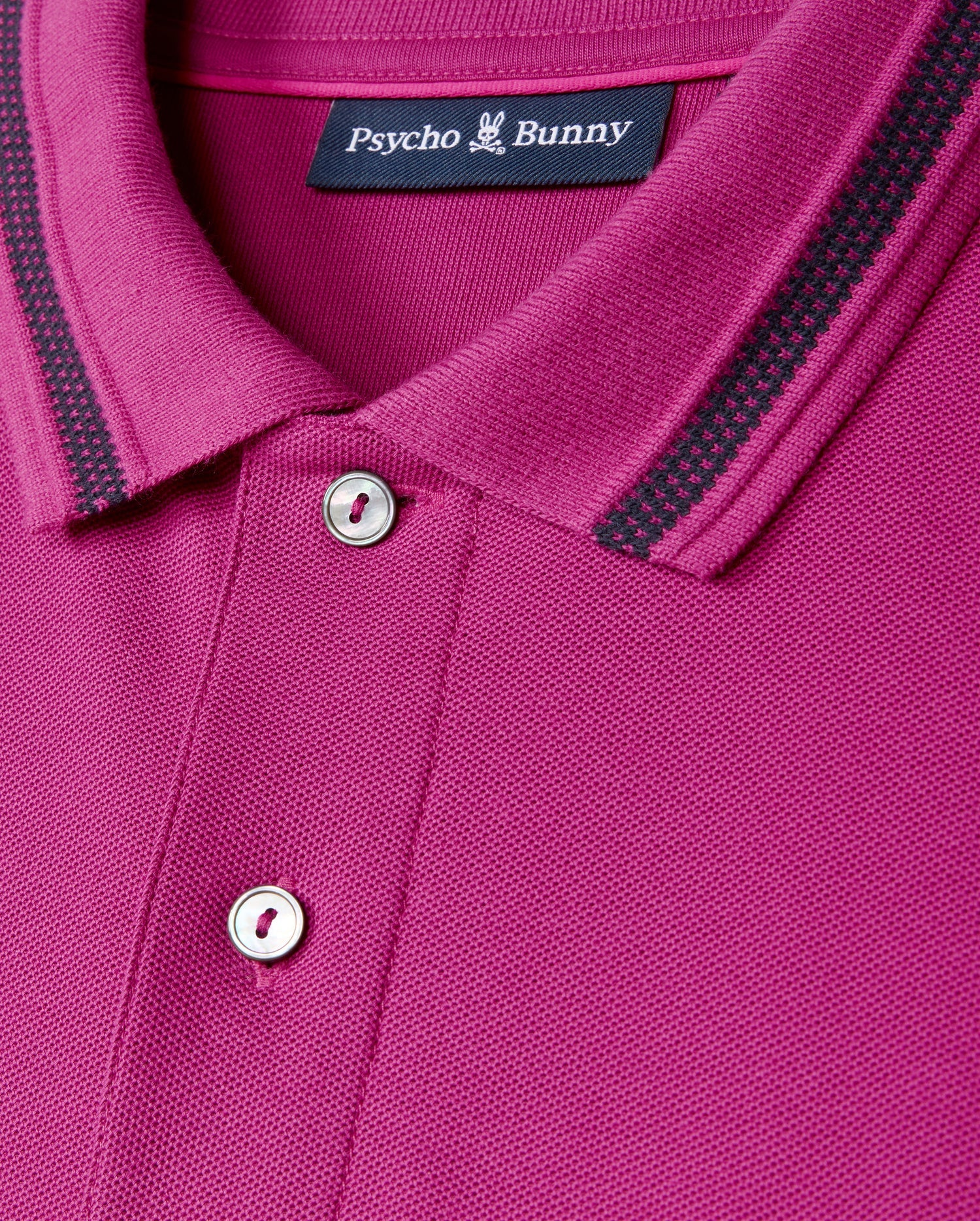 Close-up of the MENS RIVIERA PIQUE POLO SHIRT by Psycho Bunny in a vibrant magenta Pima cotton, featuring a collar with black and gray striped details. The shirt boasts two buttons, an embroidered Bunny appliqué, and a label at the collar that reads "Psycho Bunny" with a small bunny logo.