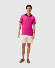 A man stands against a light gray background, wearing a bright pink Psycho Bunny Richards Johnny Collar Pique Polo Shirt (B6K641C200) with a navy collar made of breathable Pima cotton. He pairs it with beige shorts and brown loafers. His short curly hair complements his slight smile, and he keeps his arms relaxed by his sides.