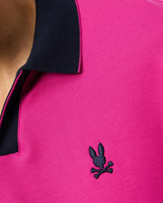 Close-up of a person wearing the Psycho Bunny Men's Richards Johnny Collar Pique Polo Shirt (B6K641C200) in pink, featuring a contrasting black collar. This breathable Pima cotton polo showcases an embroidered black logo on the left side of the chest, depicting a rabbit head with crossed bones. Part of the person's neck and shoulder are visible.