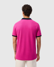Man with short, curly hair wearing a bright pink Psycho Bunny Mens Richards Johnny Collar Pique Polo Shirt - B6K641C200 made of breathable Pima cotton, featuring navy collar and sleeve cuffs, and light-colored shorts. He has his back to the camera, displaying the solid pattern design of the shirt against a plain, light background.