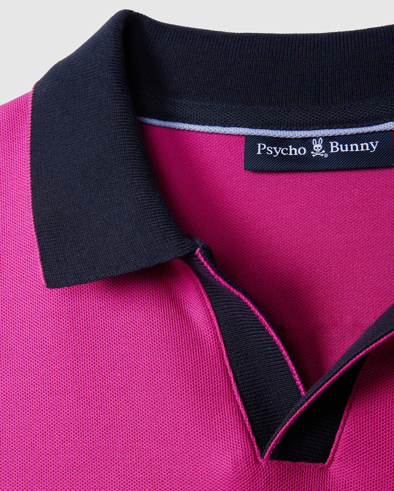 Close-up of a magenta collared Psycho Bunny MENS RICHARDS JOHNNY COLLAR PIQUE POLO SHIRT - B6K641C200 with black trim around the collar. Made from breathable Pima cotton, the inside label reads "Psycho Bunny" with a small bunny logo above the text. The solid pattern stands out against the plain light gray background.