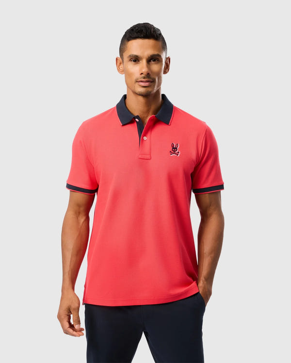 A man stands confidently wearing a red MENS KAYDEN PIQUE POLO - B6K675C200 by Psycho Bunny with black trim on the collar and sleeves. The shirt, made of diamond-knit Pima cotton, features a black emblem on the left side of the chest. He has short, dark hair and is wearing black pants. The background is plain white.