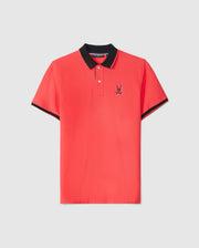 A red MENS KAYDEN PIQUE POLO - B6K675C200 by Psycho Bunny with a black collar and black-trimmed sleeves. The shirt features a small embroidered design of a bunny skull and crossbones on the left chest area. Made from diamond-knit Pima cotton, it has two buttons on the placket, giving it an overall sporty and casual look.