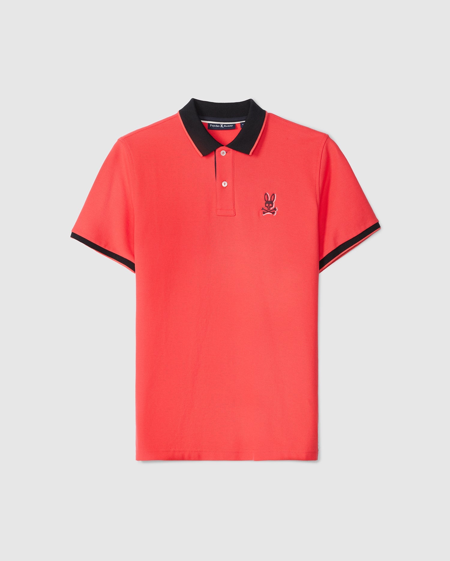 A red MENS KAYDEN PIQUE POLO - B6K675C200 by Psycho Bunny with a black collar and black-trimmed sleeves. The shirt features a small embroidered design of a bunny skull and crossbones on the left chest area. Made from diamond-knit Pima cotton, it has two buttons on the placket, giving it an overall sporty and casual look.