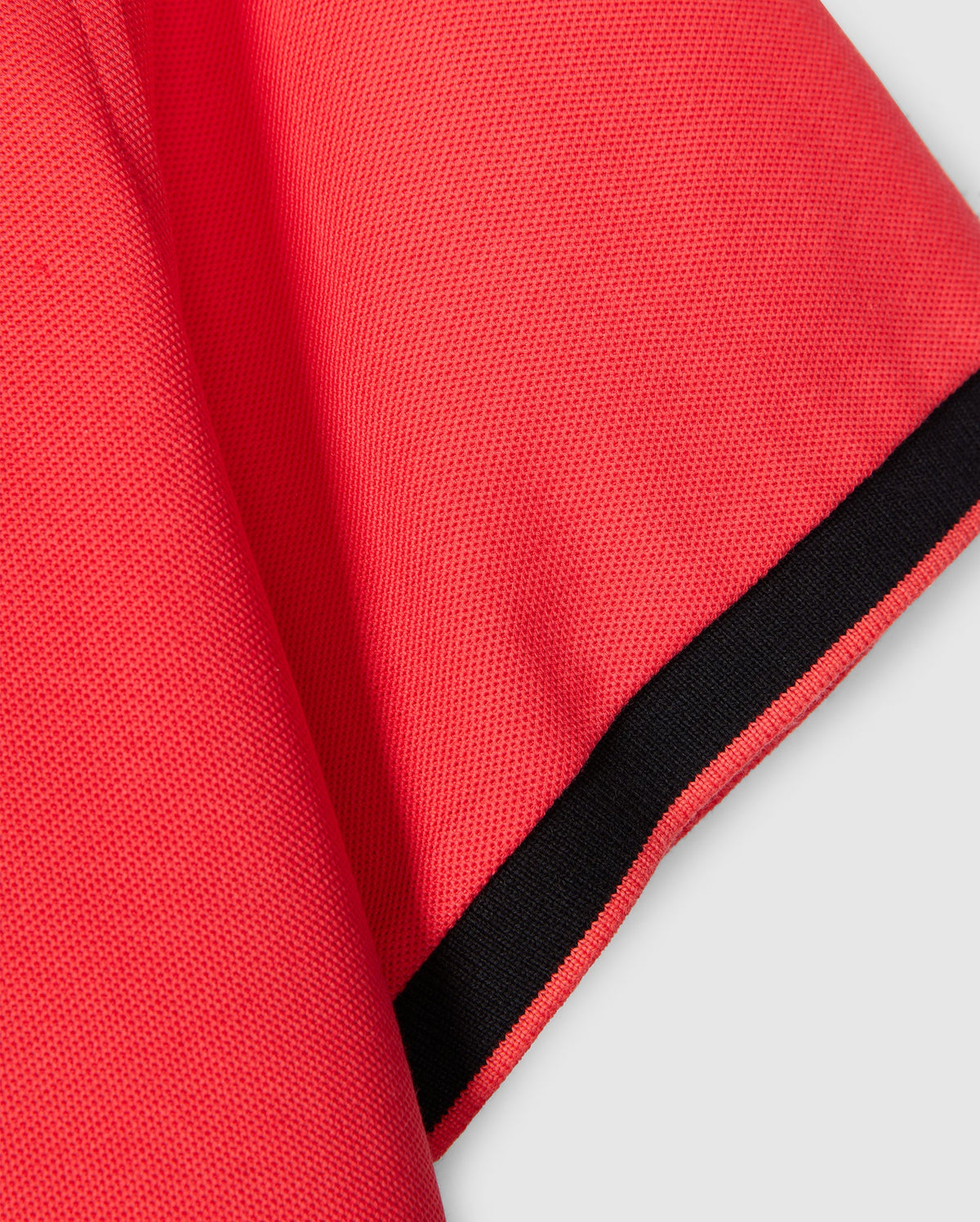 Close-up of a red fabric sleeve with a small section of the shoulder visible. The sleeve has black edging at the hem, adding contrast to the texture and color of the diamond-knit Pima cotton MENS KAYDEN PIQUE POLO - B6K675C200 by Psycho Bunny. The background is a neutral gray.
