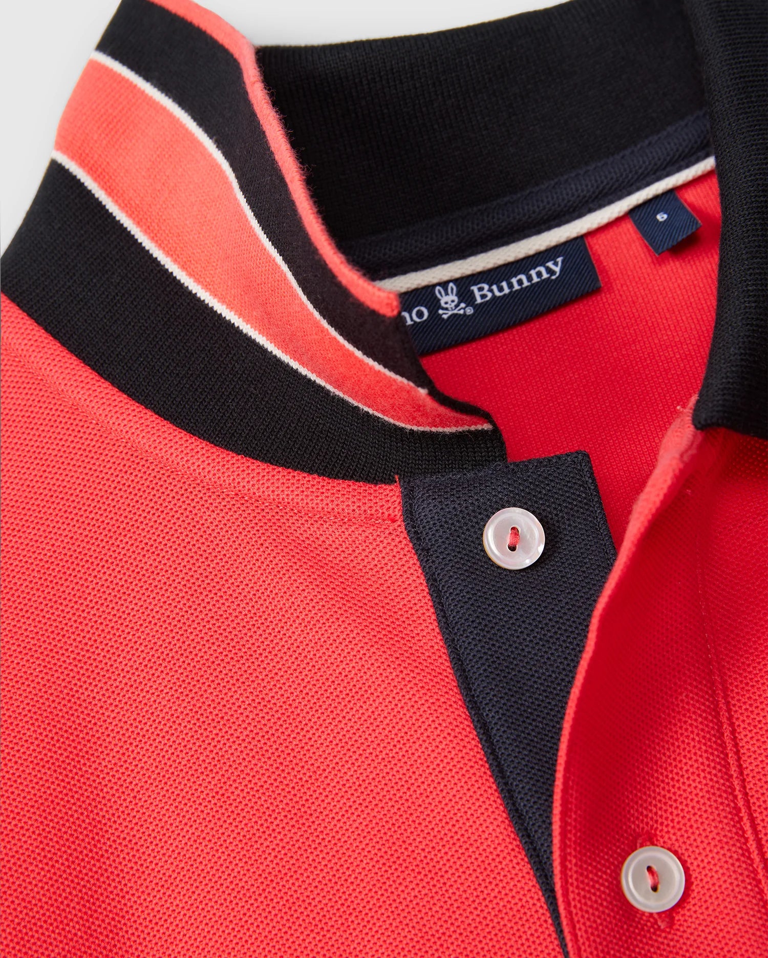 Close-up of a red MENS KAYDEN PIQUE POLO - B6K675C200 with black and white contrasting stripes on the collar and button placket. The brand label inside the collar reads "Psycho Bunny" and features a small bunny logo. Made from diamond-knit Pima cotton, the shirt is neatly folded at the collar.