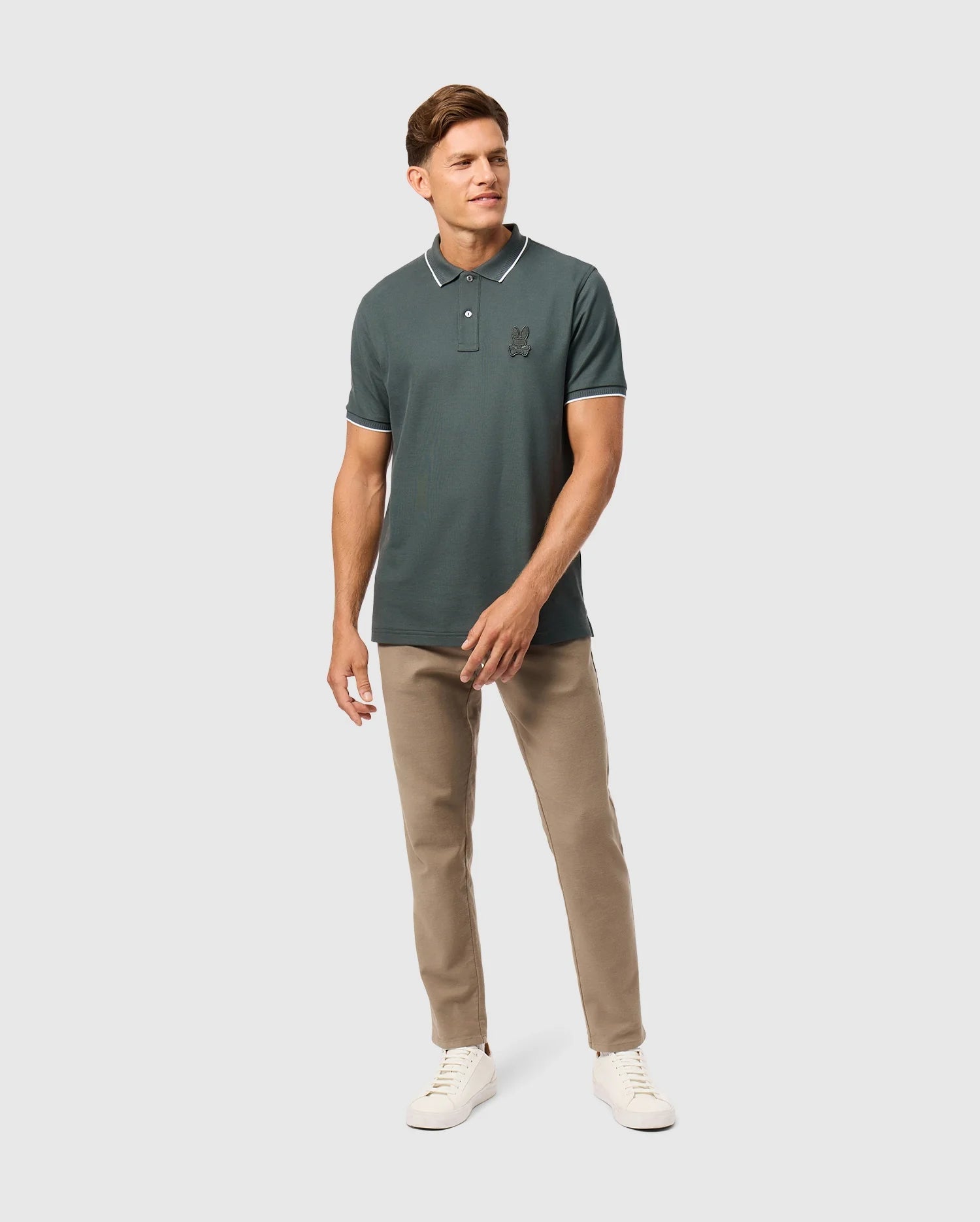 A man stands confidently against a plain background, wearing a dark green Mens Kingston Pique Polo Shirt (B6K764D200) by Psycho Bunny, featuring an embroidered Bunny patch on the chest. He pairs it with beige pants and white sneakers. The Pima cotton fabric of the polo shirt adds a touch of casual elegance. He has short hair and shows a slight smile, with his left hand relaxed at his side and his right hand slightly bent.