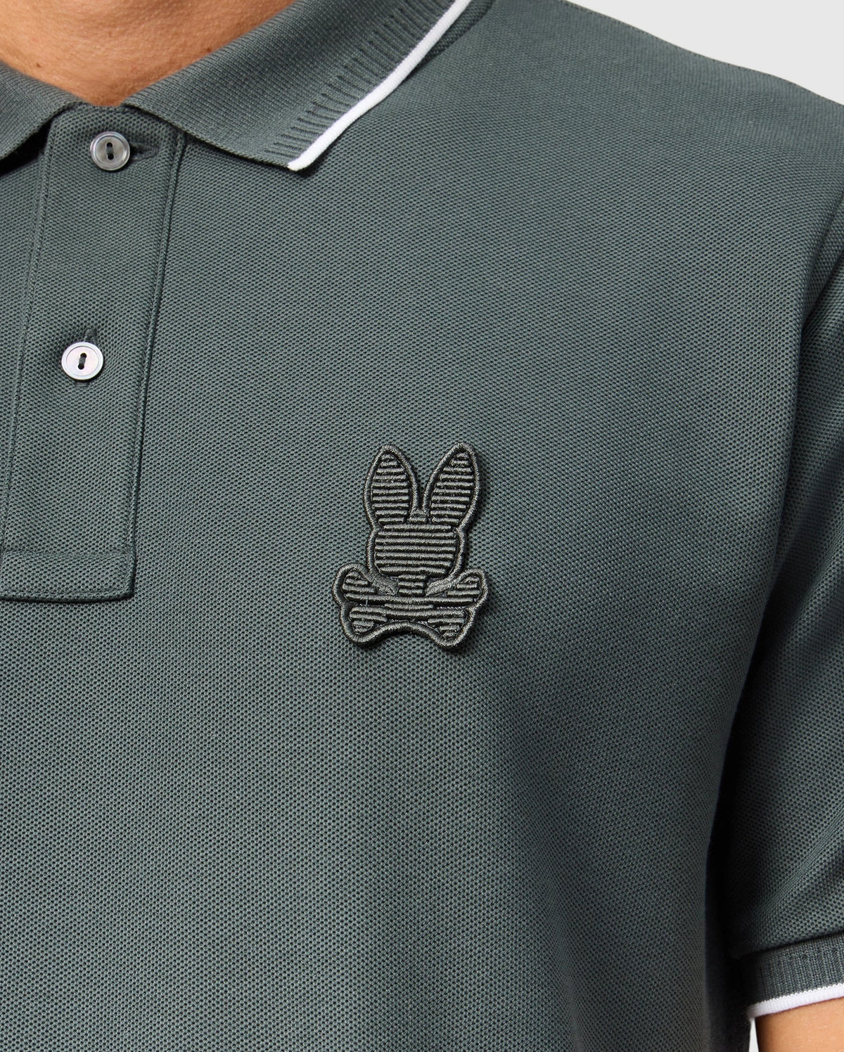 A close-up of a person wearing a green Psycho Bunny Men's Kingston Pique Polo Shirt (B6K764D200), made from Pima cotton, featuring an embroidered Bunny patch logo on the left side of the chest. The shirt has a two-button placket and white trim on the collar. Only the upper torso is visible.