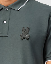 A close-up of a person wearing a green Psycho Bunny Men's Kingston Pique Polo Shirt (B6K764D200), made from Pima cotton, featuring an embroidered Bunny patch logo on the left side of the chest. The shirt has a two-button placket and white trim on the collar. Only the upper torso is visible.
