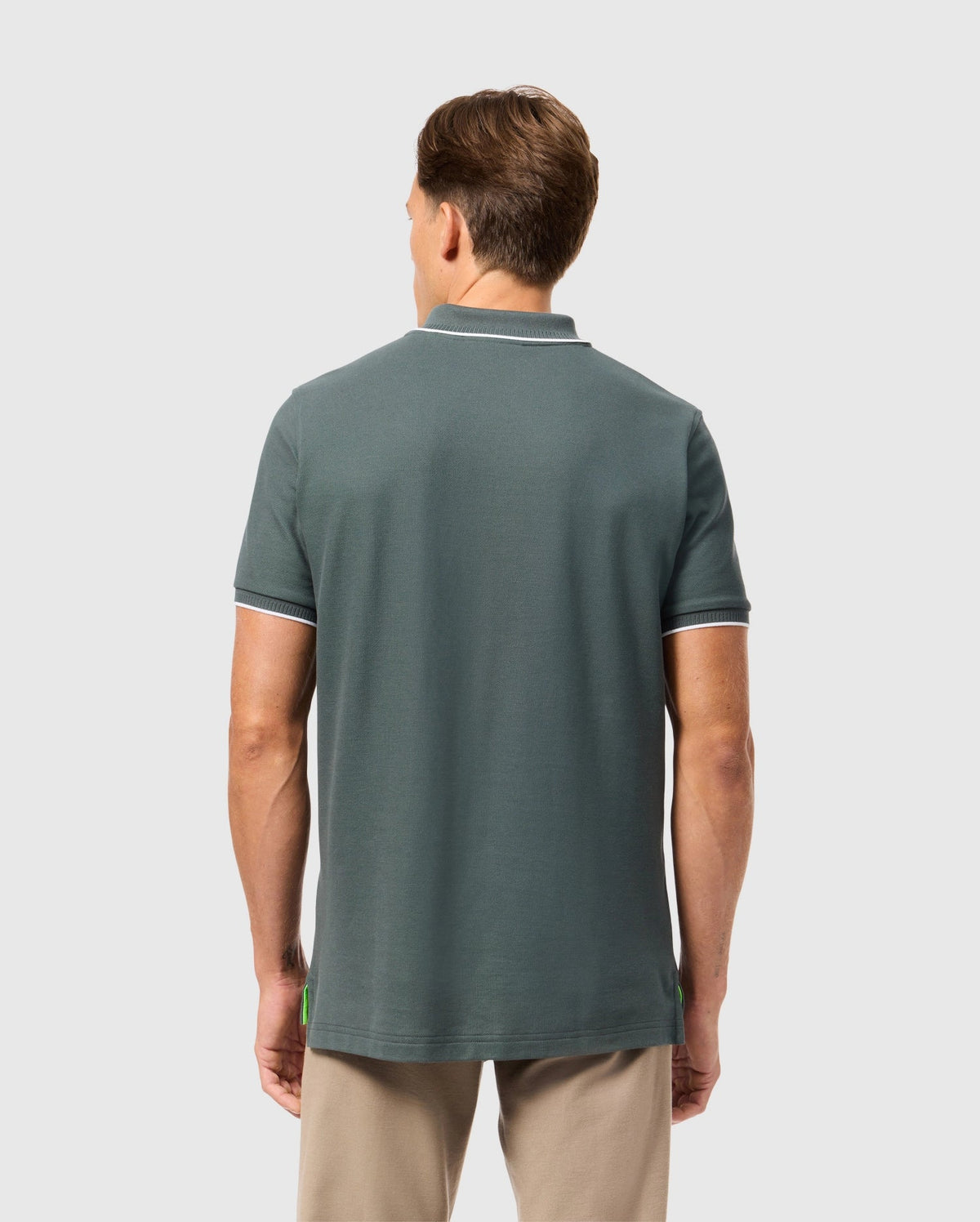 A man with short hair is standing with his back to the camera, wearing a green Psycho Bunny MENS KINGSTON PIQUE POLO SHIRT - B6K764D200 made of Pima cotton and beige pants. The plain, light-colored background highlights his casual attire and the embroidered Bunny patch on his shirt.