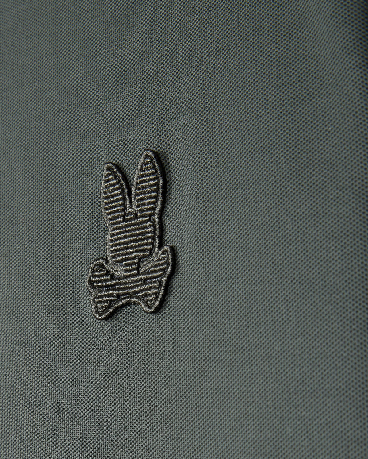 A close-up view of the Psycho Bunny Men's Kingston Pique Polo Shirt (B6K764D200) showcases an embroidered Bunny patch featuring a striped bunny head above two crossed bones, reminiscent of the Jolly Roger flag. The design is stitched with darker thread that subtly contrasts with the fabric.
