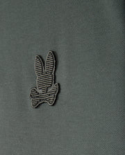 A close-up view of the Psycho Bunny Men's Kingston Pique Polo Shirt (B6K764D200) showcases an embroidered Bunny patch featuring a striped bunny head above two crossed bones, reminiscent of the Jolly Roger flag. The design is stitched with darker thread that subtly contrasts with the fabric.