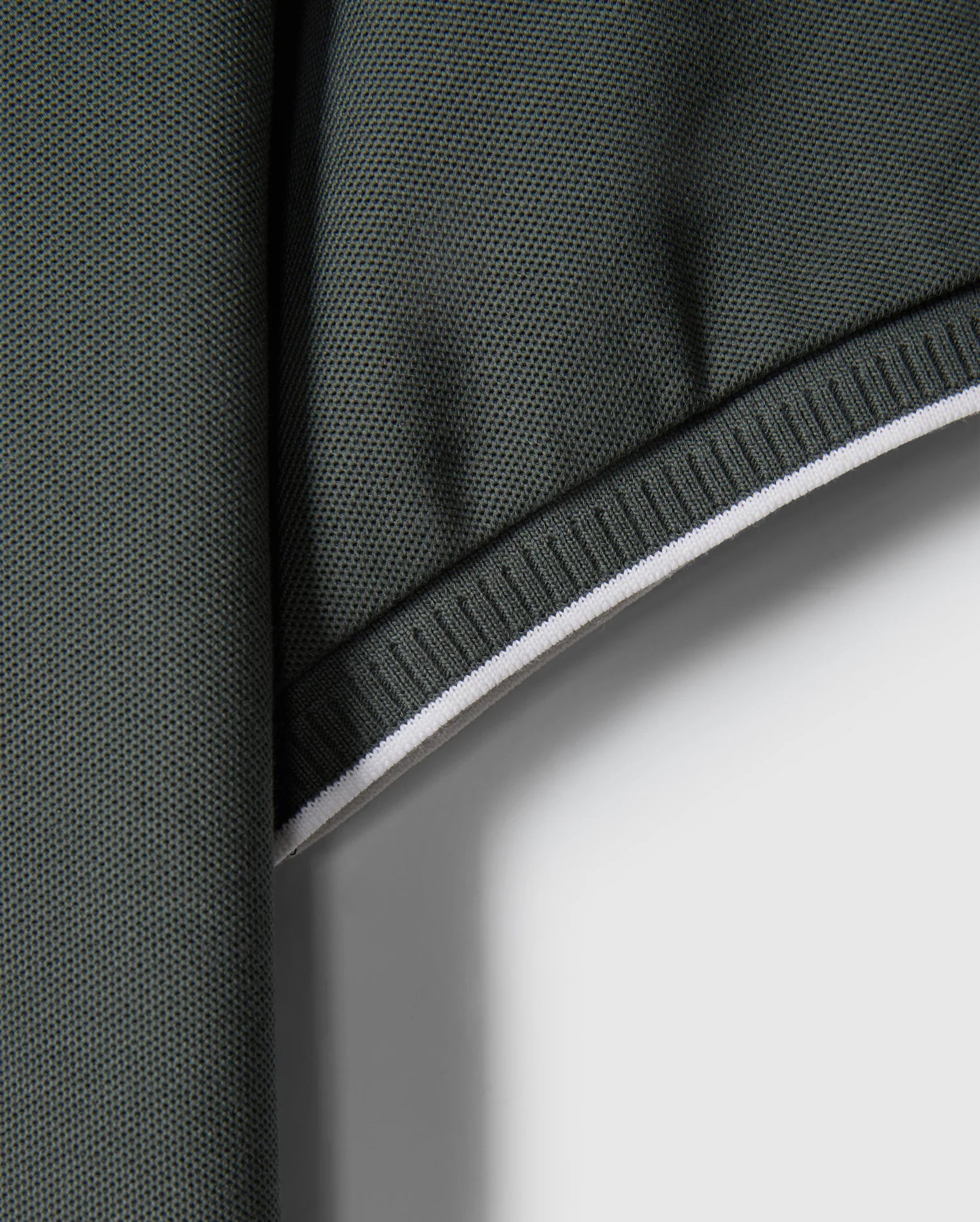 Close-up of a green Pima cotton fabric with a textured surface and white edge. The fabric appears to be folded at the top, creating a shadowed area. The edge is reinforced with white stitching, providing contrast against the dark green material – perfect for crafting a stylish MENS KINGSTON PIQUE POLO SHIRT - B6K764D200 by Psycho Bunny, perhaps with an embroidered Bunny patch.