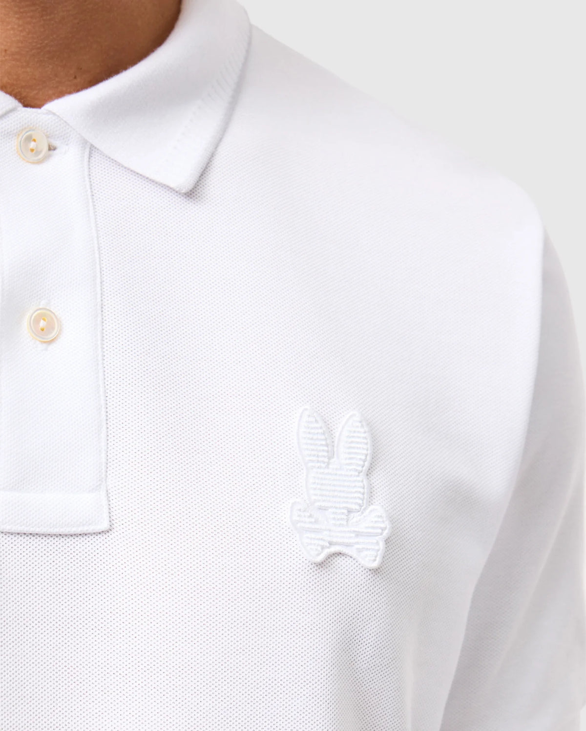 Close-up of a person wearing a white Psycho Bunny MENS KINGSTON PIQUE POLO SHIRT - B6K764D200, made from Pima cotton, featuring button details and an embroidered Bunny patch with crossed bones on the left side of the chest. The background is a neutral gray.