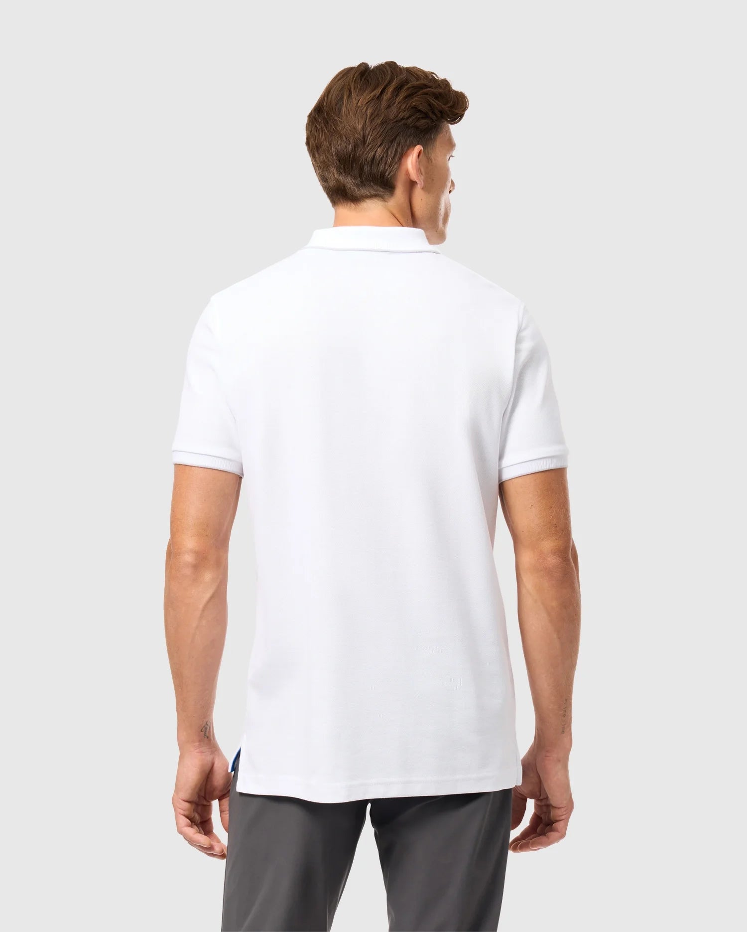 A man with short brown hair is facing away from the camera, wearing a white Psycho Bunny Men's Kingston Pique Polo Shirt (B6K764D200) with an embroidered Bunny patch, along with grey pants. The background is plain and light-colored.