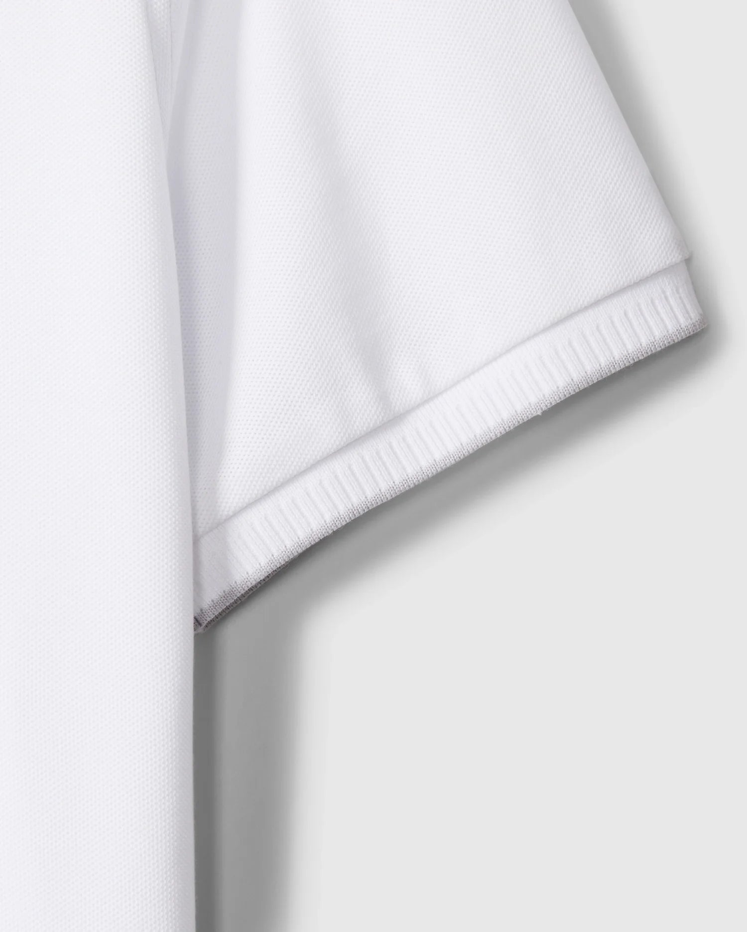 A close-up of the sleeve on the MENS KINGSTON PIQUE POLO SHIRT - B6K764D200 by Psycho Bunny highlights the textured Pima cotton fabric and detailed hem stitching. The white, short-sleeved polo shirt is crafted from light, breathable material. An embroidered Bunny patch adds a touch of charm against the flat, neutral gray background.