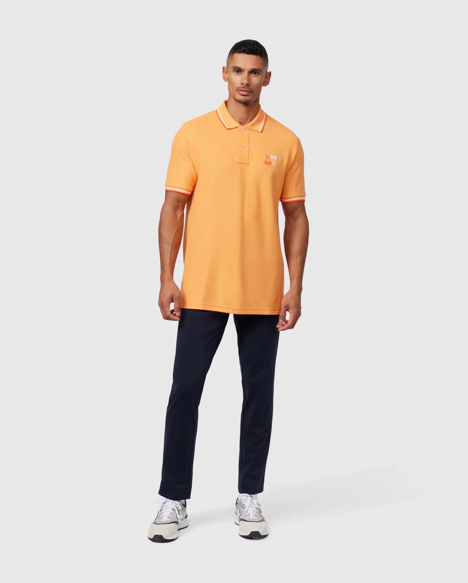 A man is standing against a plain background wearing an orange Psycho Bunny MENS WINTON PIQUE POLO SHIRT - B6K841A2PC with a small embroidered emblem on the left chest, navy blue pants, and white sneakers. Made from Peruvian Pima cotton in a diamond-knit piqué, the polo complements his look perfectly. He has short hair and is looking directly at the camera.