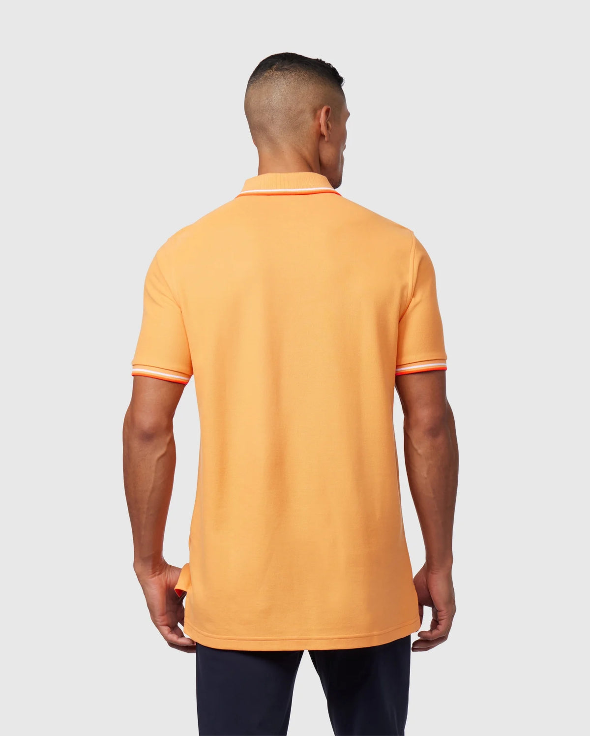 A man with short hair is facing away from the camera, wearing an orange Psycho Bunny MENS WINTON PIQUE POLO SHIRT - B6K841A2PC made of Peruvian Pima cotton, featuring white and orange striped trim on the sleeves and collar. He has on dark pants and stands against a plain light gray background.