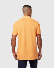 A man with short hair is facing away from the camera, wearing an orange Psycho Bunny MENS WINTON PIQUE POLO SHIRT - B6K841A2PC made of Peruvian Pima cotton, featuring white and orange striped trim on the sleeves and collar. He has on dark pants and stands against a plain light gray background.
