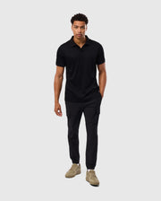 The individual is positioned against a plain background, modeling the Psycho Bunny MENS BROADWAY PIMA INTERLOCK JOHNNY COLLAR POLO - B6K891D200, crafted from mercerized Pima cotton. They pair this top with black cargo pants, one hand casually placed in a pocket, and finish the look with light brown sneakers.