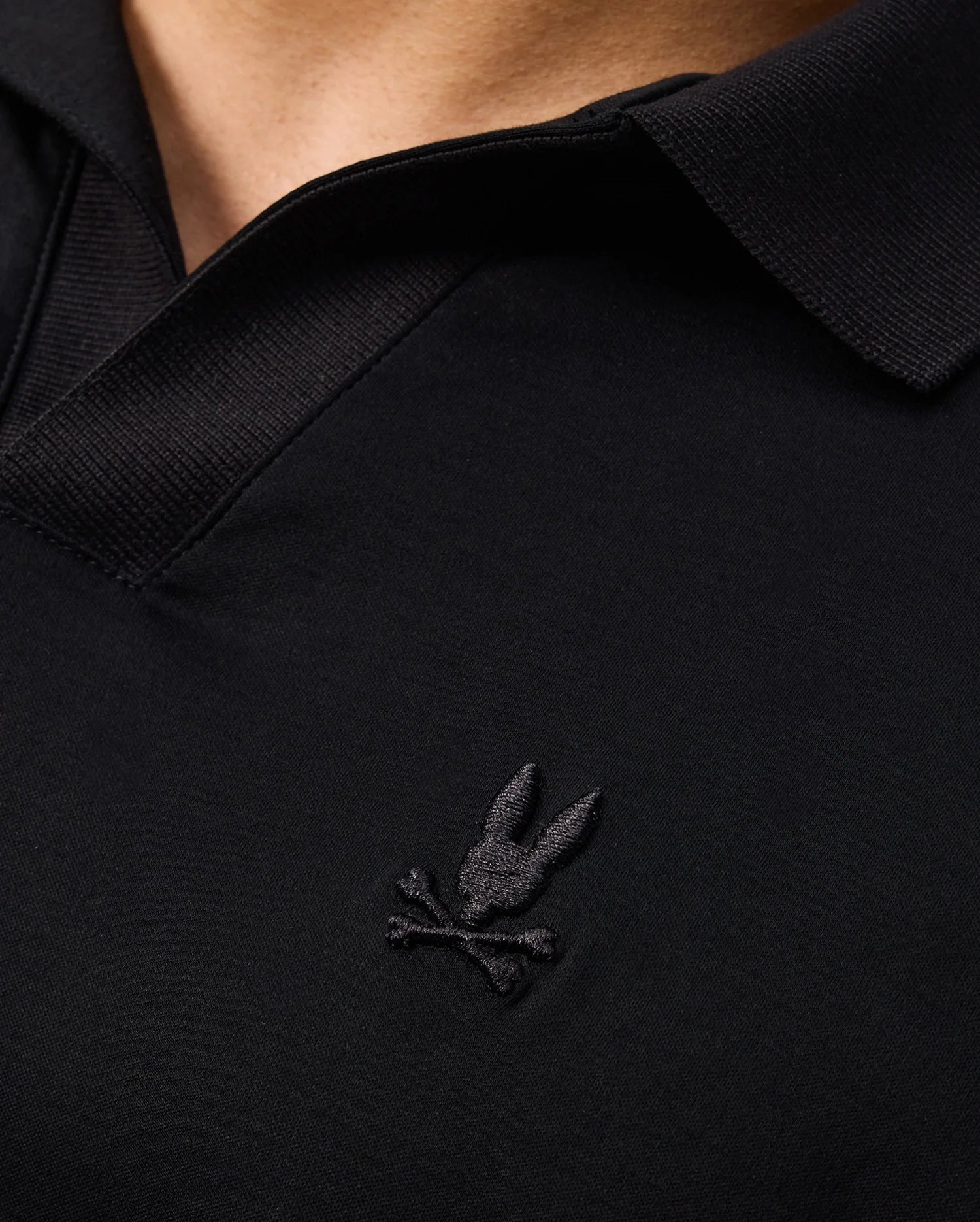Close-up of a black MENS BROADWAY PIMA INTERLOCK JOHNNY COLLAR POLO by Psycho Bunny, crafted from durable lustre mercerized Pima cotton and featuring a stitched logo with a rabbit head above crossed bones. The shirt's collar and part of the neckline are visible.