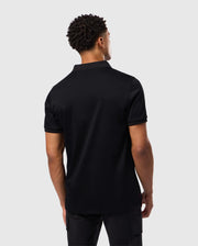 A man faces away from the camera, dressed in a Psycho Bunny MENS BROADWAY PIMA INTERLOCK JOHNNY COLLAR POLO in black. His black pants complete the ensemble, and the light gray background accentuates the durable luster of his mercerized Pima cotton apparel.